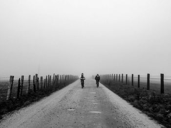 Rear view of people riding bicycle on dirt road against clear sky