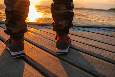 Low section of man standing on wooden pier against sky during sunset