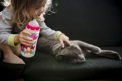 Toddler girl patting her grey cat pet while drinking milk outdoor