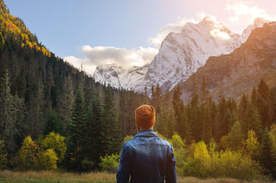 Back view of a man in a knitted hat and denim jacket against the backdrop of a green dense forest