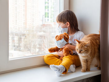 Toddler boy sits with teddy bear, both in medical masks. sick kid with cute ginger cat. 