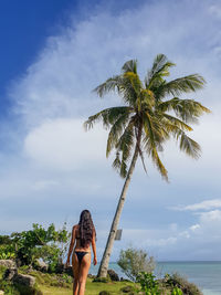 Rear view of woman in bikini standing at beach against cloudy sky