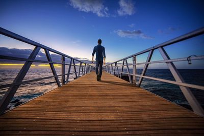 Rear view of mature man walking on pier over sea against sky during sunset