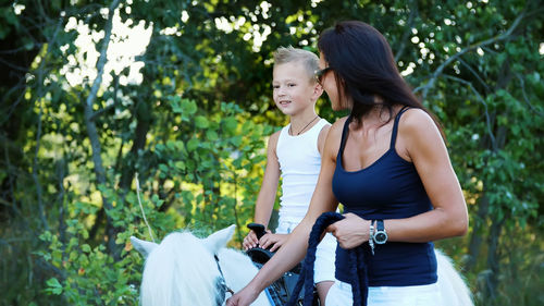 A woman and a boy are walking around the field, son is riding a pony, mother is holding a pony for