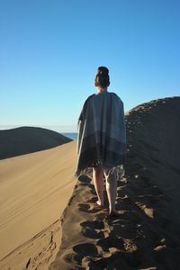 Rear view of woman with blanket walking on sand dune in desert against clear sky