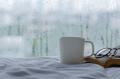 A cup of coffee with book and spectacles on bed with rain drop on window. stay home and relaxing.