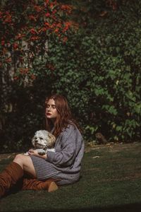 Young woman holding dog while sitting on grassy field against trees