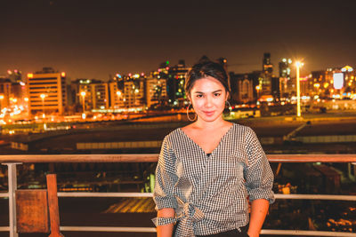 Portrait of smiling young woman standing against illuminated cityscape at night