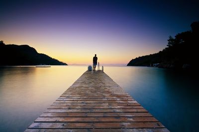 Rear view of man standing on pier at lake against clear sky during sunset