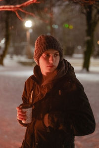 A young man in a gloomy winter forest, in a knitted warm hat, snow around, holding a warming drink 