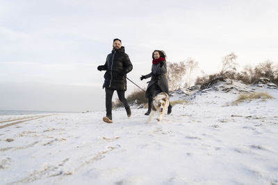 Young couple running with dog on snow at sunset