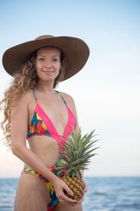 Portrait of young woman wearing hat and holding pineapple against sky