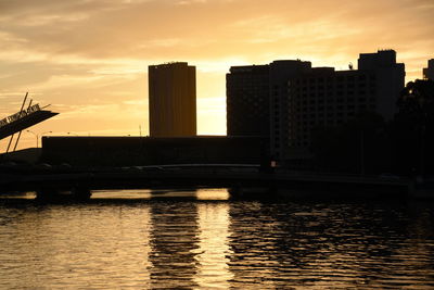 Silhouette buildings by river against sky during sunset