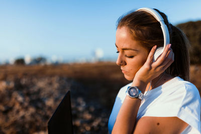 Young woman wearing headphones while using laptop against sky