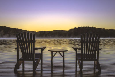 Chairs by lake against clear sky during sunset