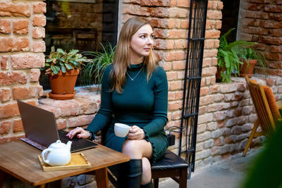 Woman holding coffee cup while using laptop on table at sidewalk cafe