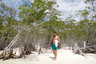 Shirtless man walking on sand against trees at beach