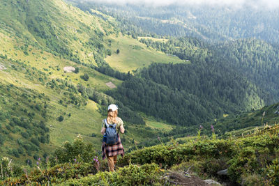 Rear view of young woman with backpack hiking in mountains among rhododendron plants and flowers 