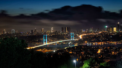 Illuminated bosphorus bridge in city against cloudy sky at night