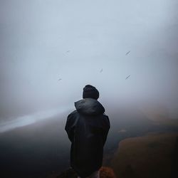 Rear view of man standing on mountain against sky during foggy weather