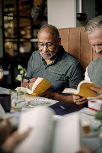 Senior man wearing eyeglasses reading book while sitting by male friend in cafe