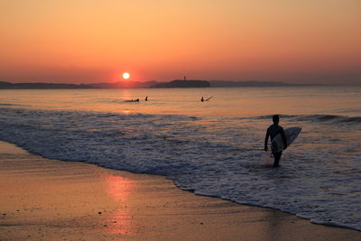 Man with surfboard standing on beach against sky during sunset