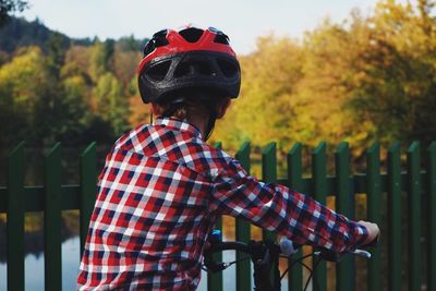 Rear view of boy wearing helmet with bicycle standing by fence against lake at park