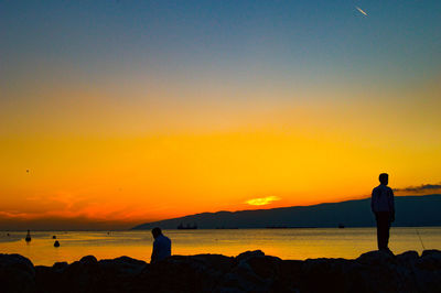 People standing on rock by sea against sky during sunset