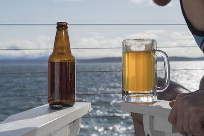 Cropped image of man with beer glass while sitting on chair by sea