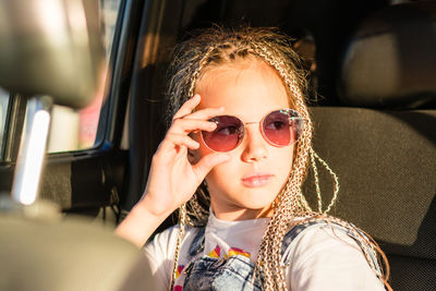 Serious teenager girl with afro-braids holds sunglasses with her hand and looks out the window 