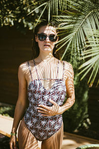 Portrait of woman wearing sunglasses in swimwear standing against trees