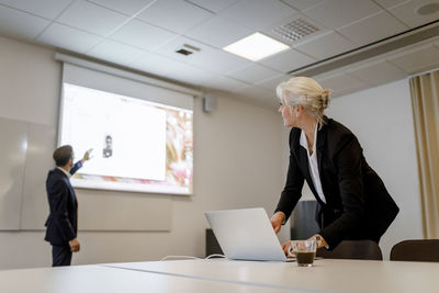 Female professional using laptop while male colleague standing by flat screen during video conference in board room at o