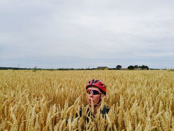 Young man wearing sunglasses and helmet sitting amidst crops at farm against cloudy sky