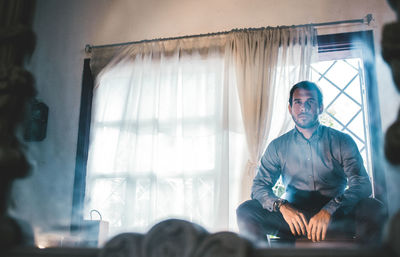 Serious man sitting on window sill reflecting in mirror
