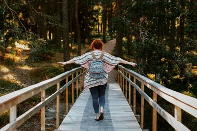 Rear view of woman with arms outstretched walking on footbridge in forest