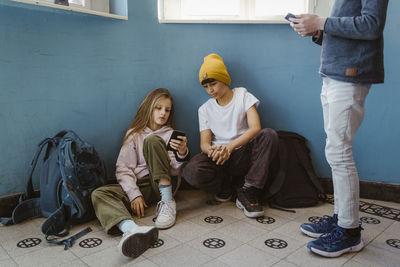 Girl sharing smart phone with male friend crouching by wall in school