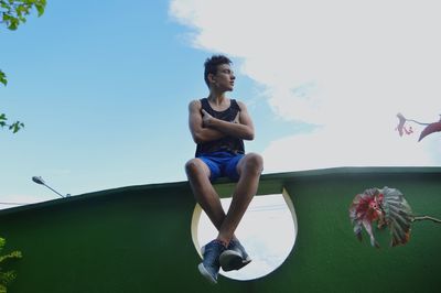 Young man sitting on surrounding wall against sky