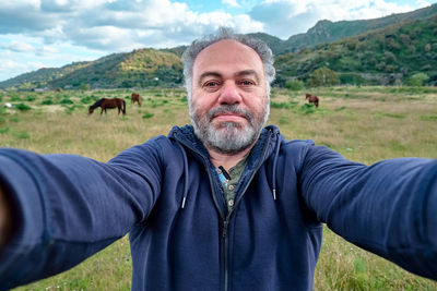 Happy, serious mature bearded man making a selfie on meadow with grazing horses.