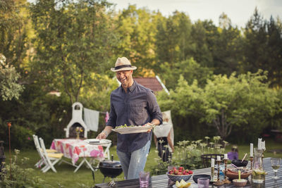 Cheerful man carrying plates by dining table in back yard during garden party