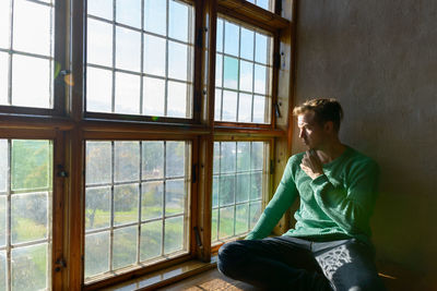 Young man looking away while sitting on window at home