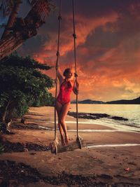Woman standing on rope swing at beach against sky during sunset