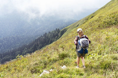 Young woman in cap and plaid shirt with big backpack hiking in green mountains against fog in summer 