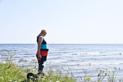 Pregnant woman standing on grass by sea against clear sky