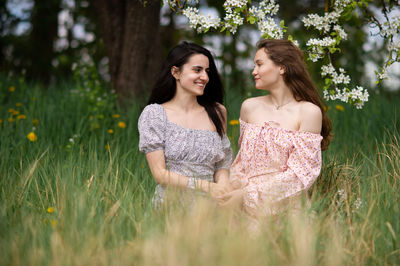 Two young girls in dresses are sitting on the green grass under a white tree and laughing