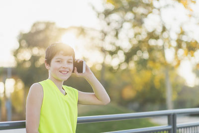 Portrait of smiling boy talking on mobile phone while standing against trees