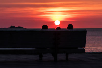 Silhouette people sitting by sea against orange sky