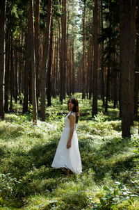 Side view of woman wearing white dress while standing on grass in forest