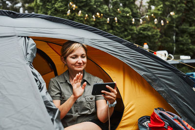 Female having video call with friends using smartphone while sitting in tent at camping. camp life