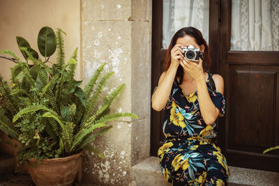 Woman photographing by potted plant outside house