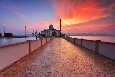 Walkway leading towards mosque by sea at sunset against sky